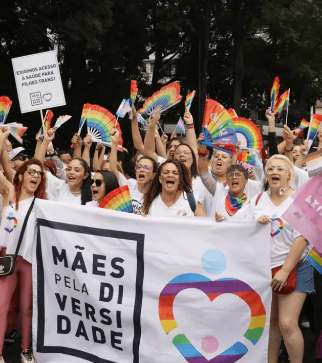 A imagem mostra um grupo de pessoas durante uma manifestação do orgulho LGBTQIAPN+, reunidas em uma marcha ao ar livre. Na frente do grupo, participantes seguram uma grande faixa branca com o nome “Mães pela Diversidade” e o símbolo de um coração colorido com as cores da bandeira LGBTQIAPN+.